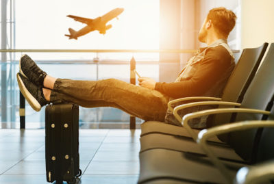 Person sitting in an airport with their phone in hand, looking out the window at a plane taking off while resting their feet on their suitcase.