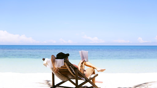Person sitting in a lounge chair on the beach, reading a book with a ball cap on.
